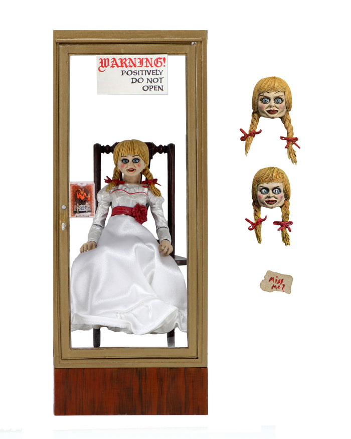 annabelle figure sitting in chair wearing white dress with hair in pig tails. interchangable heads on display. sign reading "warning! positively do not open"