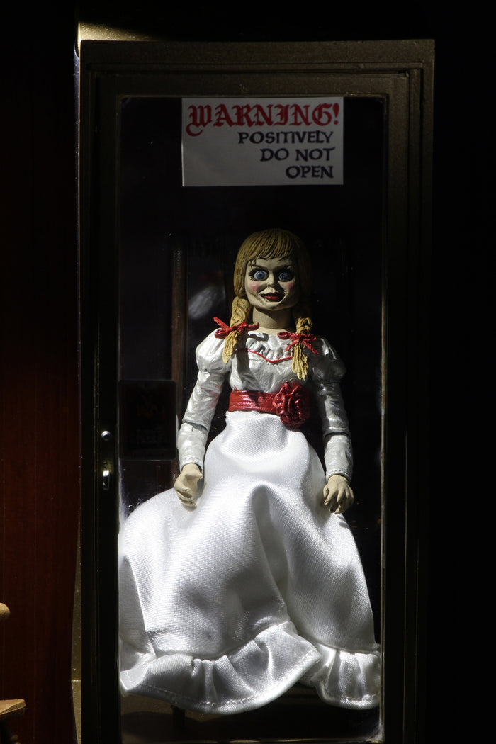 annabelle figure sitting in chair wearing white dress with hair in pig tails. sign reading "warning! positively do not open"
