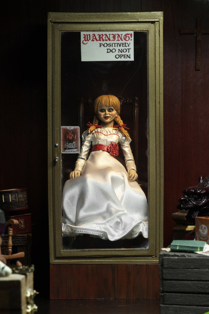 annabelle figure sitting in chair wearing white dress with hair in pig tails. sign reading "warning! positively do not open"