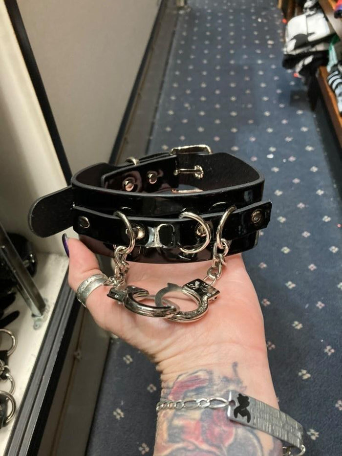Black leather collar with silver D ring details, and two silver handcuffs hanging from two silver chains.