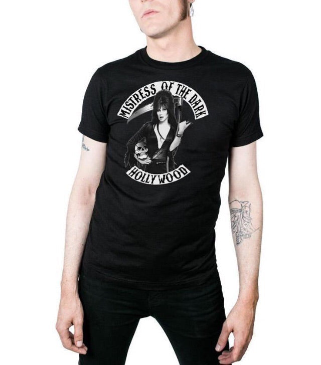 model wearing Black t-shirt with print of Elvira on front side. Print shows Elvira holding a skeleton and scythe. Top of shirt says "MISTRESS OF THE DARK" bottom says "HOLLYWOOD.