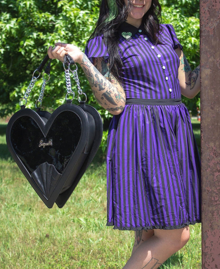 model holding heart shaped glossy black bag with a stitched spiderweb design at the bottom, chain link hardware and faux leather straps.