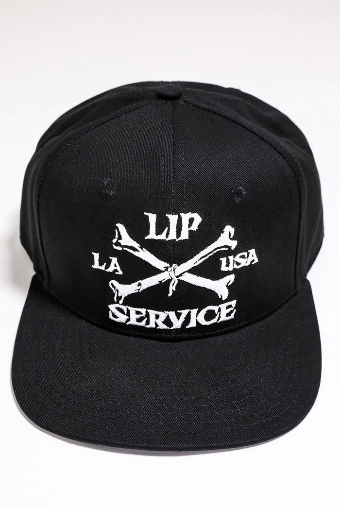 front view of Black hat with white embroidery that reads "LIP SERVICE LA USA" on the front with crossbones. Snaps on the back.