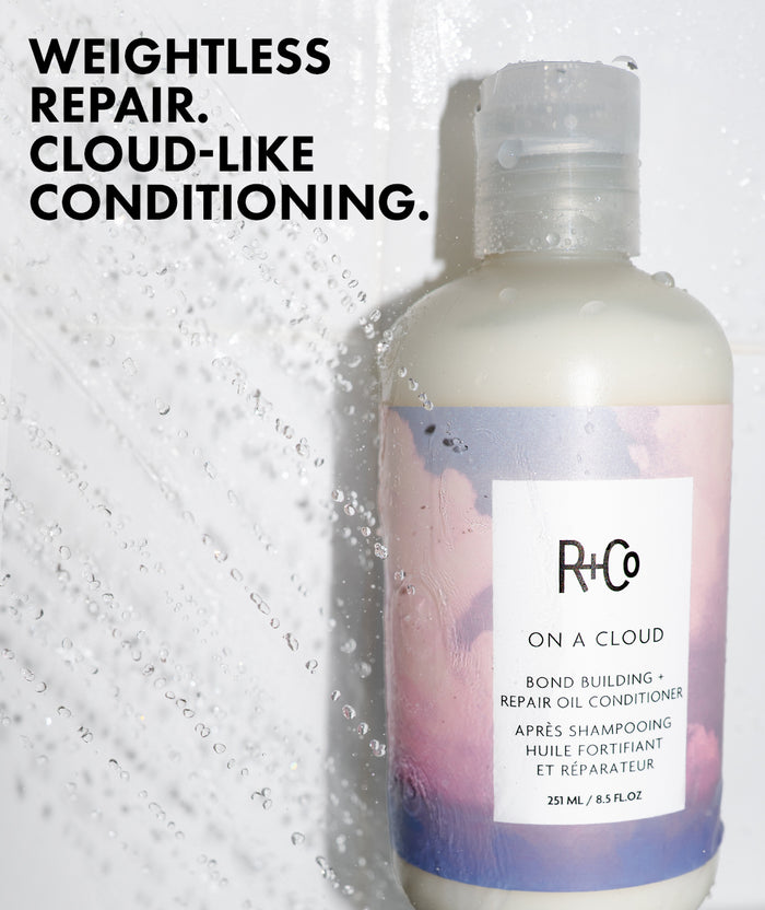 A bottle of R+Co On A Cloud Bond Building Repair Oil Conditioner shown against a white tiled background with water droplets and text reading 'WEIGHTLESS REPAIR. CLOUD-LIKE CONDITIONING.