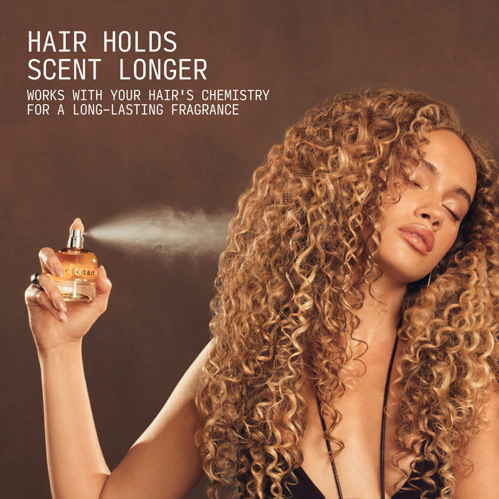 Person with long curly hair spraying Temple Oud Hair Perfume from a golden bottle onto their hair.