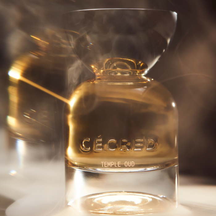 Clear glass bottle with gold liquid and "CECRED TEMPLE OUD" engraved label, surrounded by fragrant mist against warm lighting