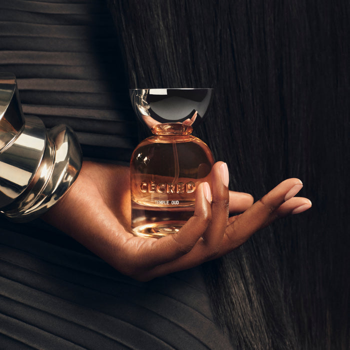 Hand holding amber glass bottle with silver cap labeled "Temple Oud" hair perfume against dark textured background.