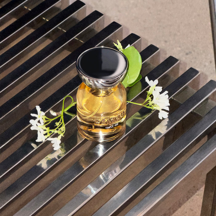 Golden hair perfume bottle with black cap surrounded by white flowers and green leaves on striped surface.