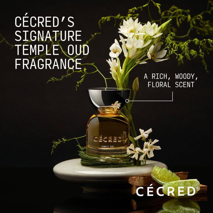 Amber glass fragrance bottle with black cap surrounded by white flowers, green foliage, and citrus on white marble surface.