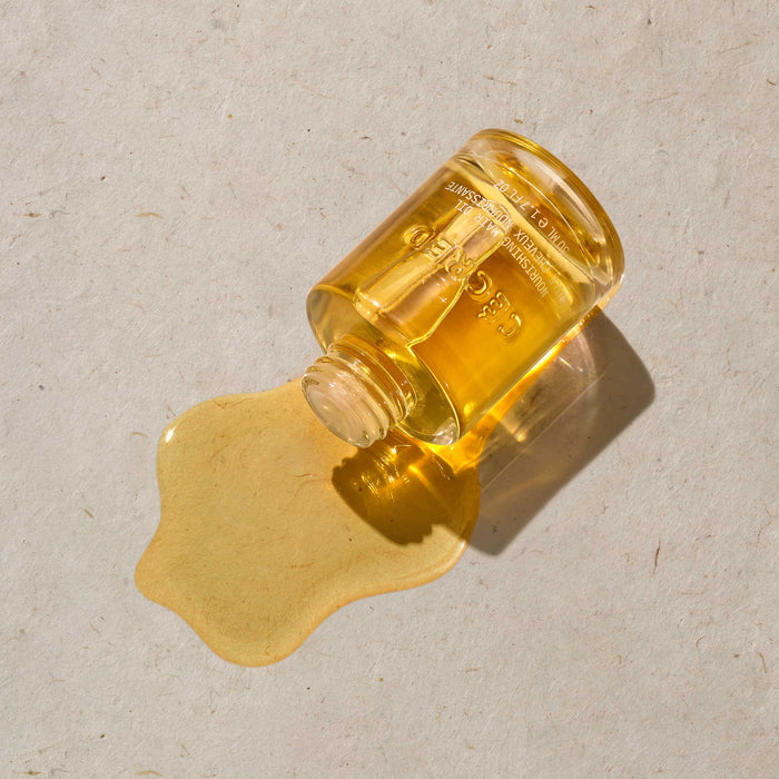 Cécred Nourishing Hair Oil amber glass bottle lying on its side with golden oil spilling out against a warm neutral textured surface