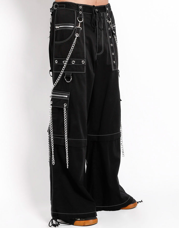 side view of Black baggy Tripp pants with white stitching. Pants have front rivet details, side pocket hanging chain details, removable attached chains, back zipper details and more. Pant legs can also zip off into shorts.