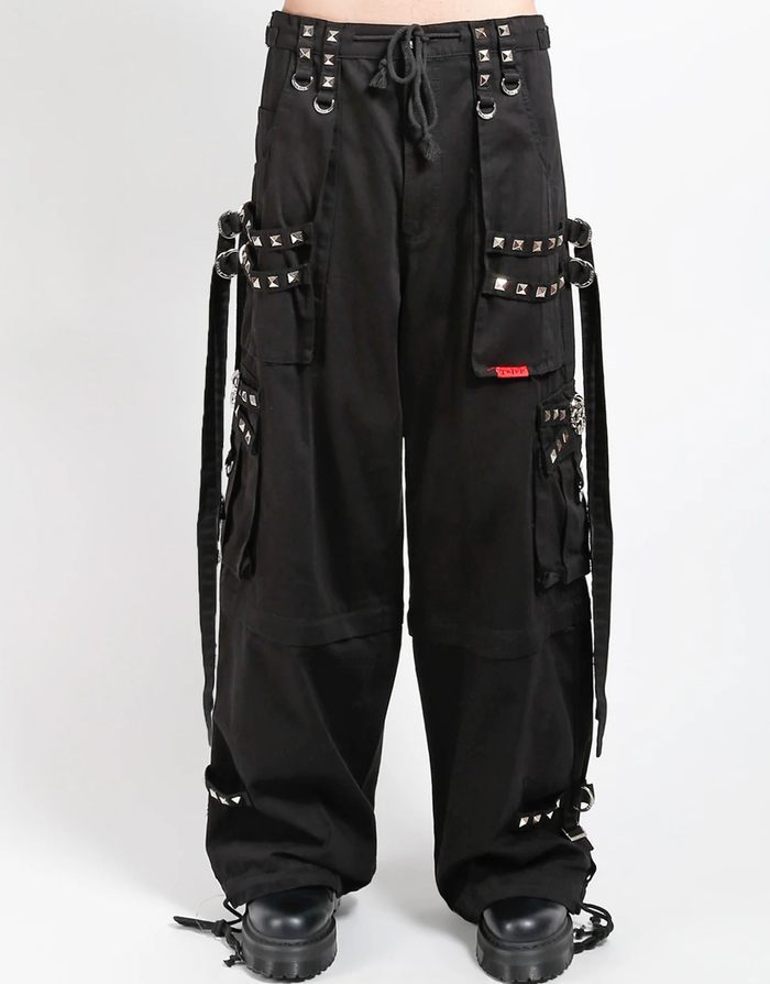 Waist-down view of baggy black punk-style cargo pants featuring silver pyramid studs, hanging side straps, a drawstring waist, and chunky black boots against a white background.