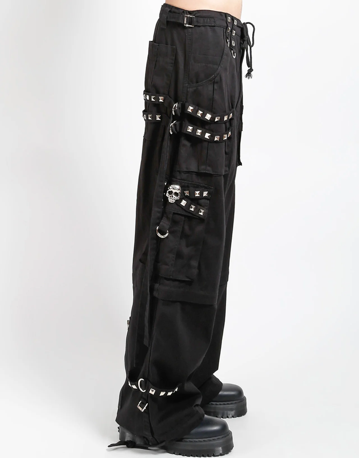 Waist-down view of baggy black punk-style cargo pants featuring silver pyramid studs, hanging side straps, a drawstring waist, and chunky black boots against a white background.