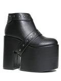 side view of black vegan leather super tall 6" heel, 4" platform sleep design boot. Boot has a rivet studded strap that wraps around and is removable or attachable with Velcro. Boot has black EVA platform.