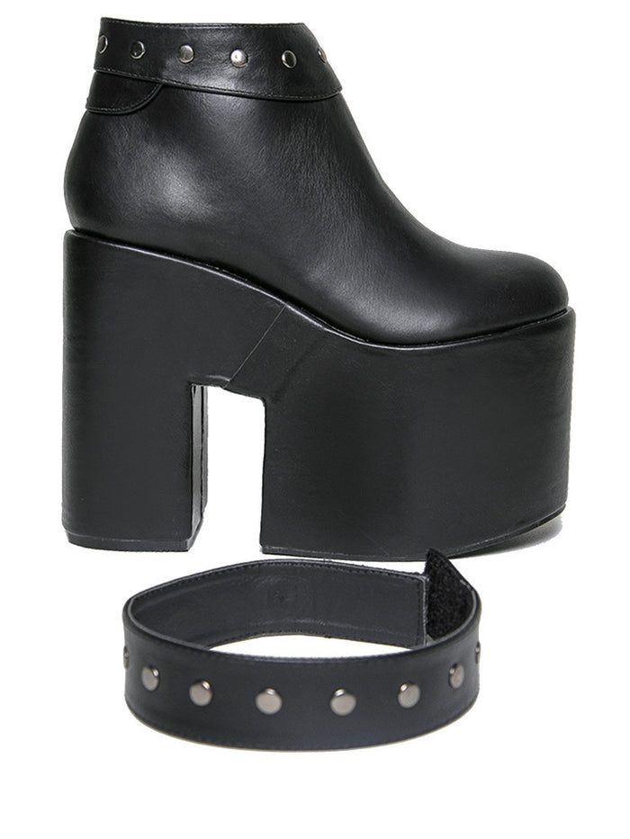 outer side view of black vegan leather super tall 6" heel, 4" platform sleep design boot. Boot has a rivet studded strap that wraps around and is removable or attachable with Velcro. Boot has black EVA platform.